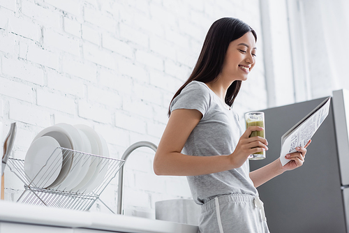 joyful asian woman with glass of fresh smoothie reading newspaper in kitchen