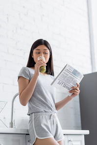 young asian woman drinking fresh smoothie while reading travel life newspaper in kitchen