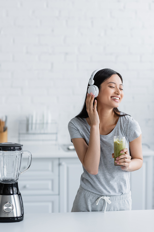 cheerful asian woman with glass of smoothie listening music in headphones near electric blender