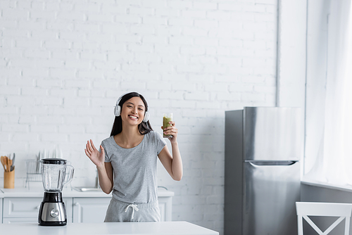 happy asian woman in headphones holding glass of smoothie and waving hand at camera near electric shaker
