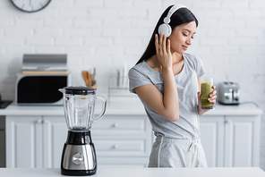 pleased asian woman with glass of smoothie listening music in headphones near electric shaker