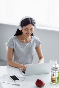 smiling asian woman in headphones typing on laptop near teapot and smartphone on desk