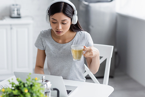 young asian woman in headphones looking at laptop while holding cup of tea