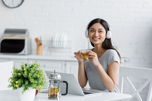 joyful asian woman in headphones holding cup of tea near laptop and blurred plant in kitchen