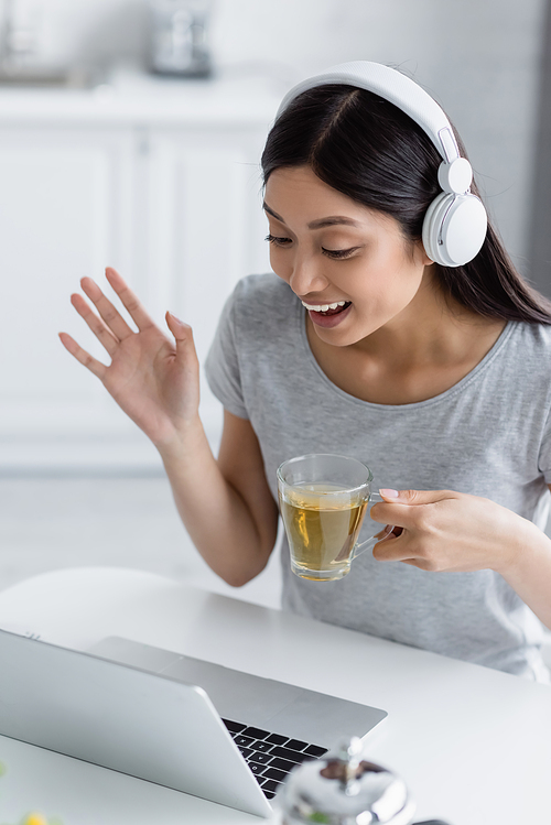excited asian woman in headphones holding cup of tea and waving hand during video call