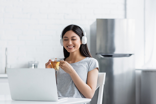 cheerful asian woman in headphones holding cup of tea while looking at laptop in kitchen