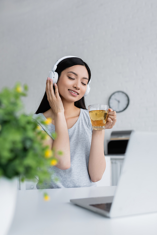 pleased asian woman relaxing in wireless headphones with cup of tea near laptop