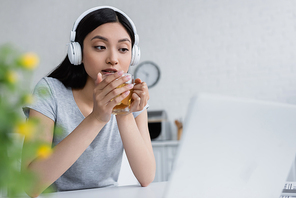 concentrated asian woman in wireless headphones holding cup of tea near laptop