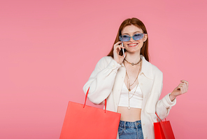 Stylish woman in sunglasses holding shopping bags and talking on smartphone isolated on pink
