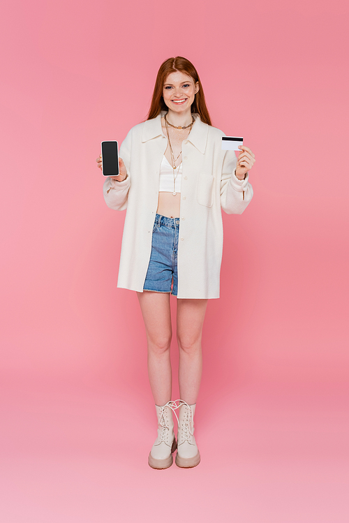 Pretty and stylish red haired woman holding cellphone and credit card on pink background