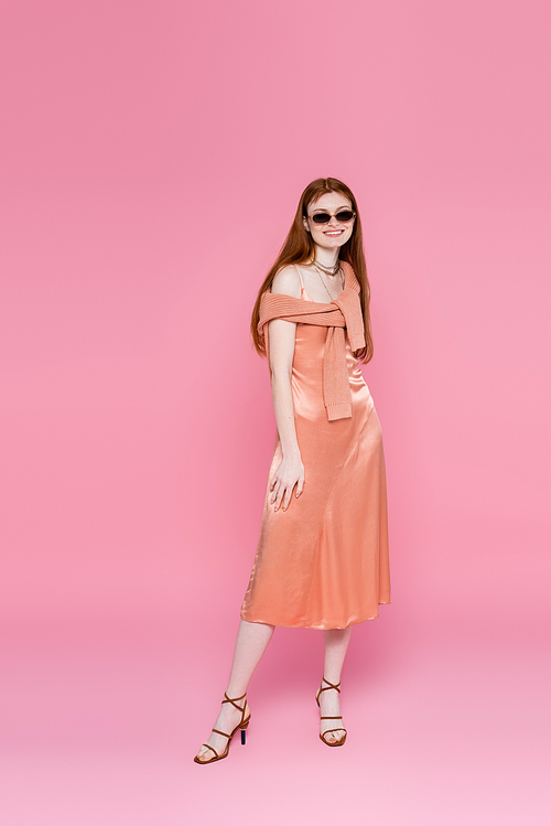 Full length of smiling woman in dress and sunglasses posing on pink background