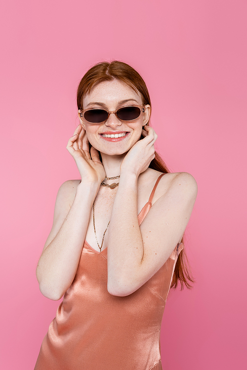 Smiling freckled woman in sunglasses and peach dress isolated on pink