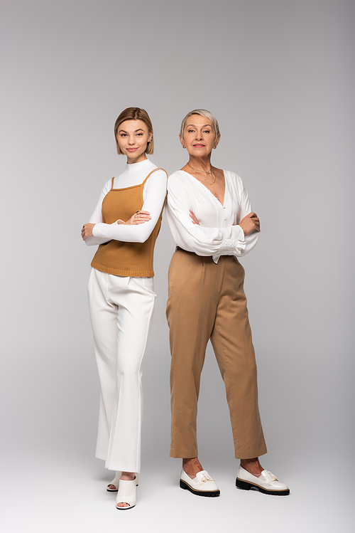 full length of cheerful middle aged mother and young daughter standing with crossed arms on grey