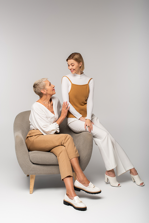 full length of happy young woman and mother looking at each other while sitting in armchair on grey