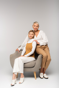 full length of positive young daughter sitting in armchair near smiling mother on grey