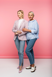 full length of stylish young woman and happy mature mother in jeans and blouse standing on pink