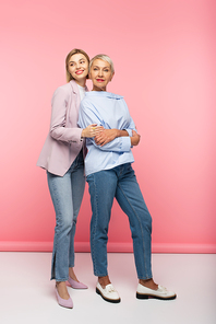 full length of happy young woman hugging mature mother in jeans and blouse while standing on pink