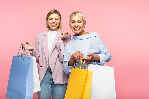 cheerful young woman and happy mature mother holding shopping bags isolated on pink