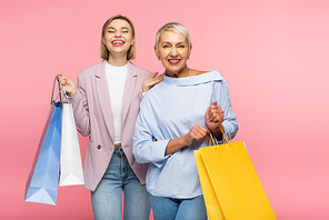 positive young woman and mature mother holding shopping bags isolated on pink