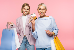 cheerful young woman and pleased mature mother holding shopping bags and credit cards isolated on pink