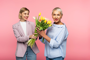 happy young daughter holding bouquet of tulips near mature mother isolated on pink