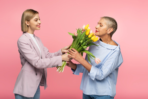pleased young daughter giving bouquet of flowers to cheerful middle aged mother isolated on pink