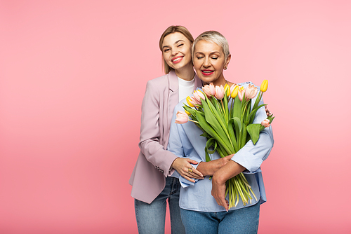 pleased daughter hugging happy middle aged mother holding bouquet of flowers isolated on pink