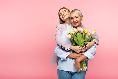 positive daughter hugging happy middle aged mother holding tulips isolated on pink