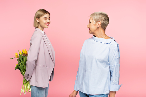 positive young daughter holding bouquet of tulips behind back near mature mother isolated on pink