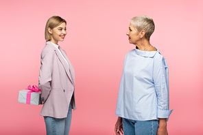 cheerful young woman holding present behind back near mature mother isolated on pink