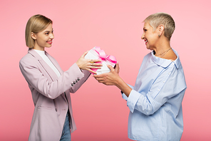 happy woman giving wrapped present to cheerful mature mother isolated on pink