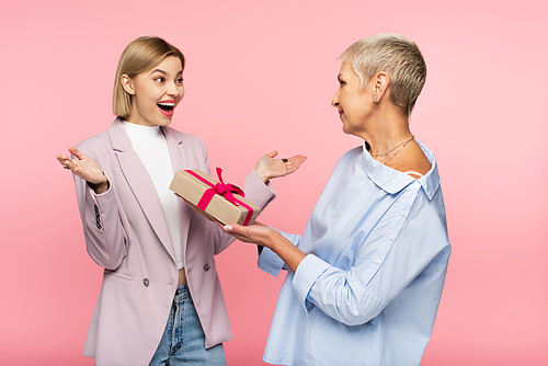 mature mother giving present to amazed daughter isolated on pink