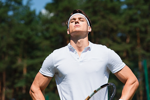 Sportsman with tennis racket standing with closed eyes outdoors