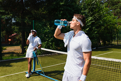 Young sportsman drinking water near african american man and blurred net on tennis court
