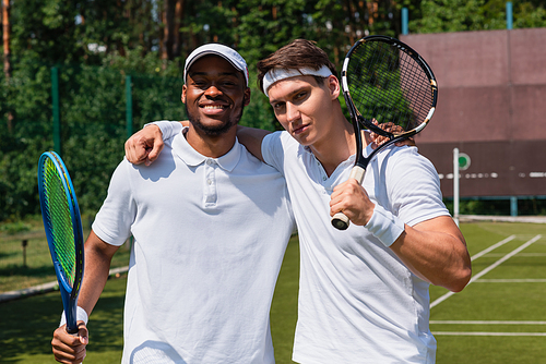 Multiethnic sportsmen with tennis rackets hugging on tennis court