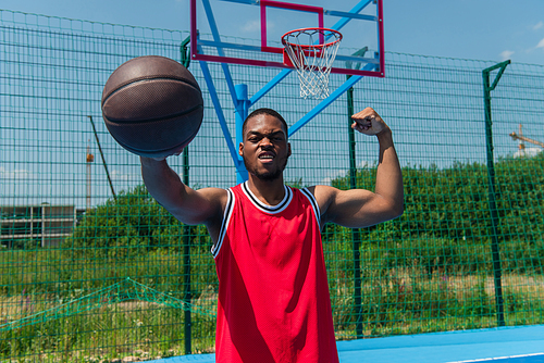 African american sportsman showing muscles and holding basketball ball on playground