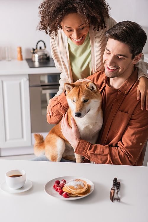 joyful man sitting with shiba inu dog near delicious pancakes and smiling african american woman