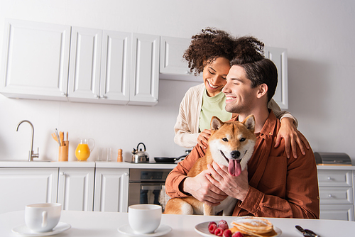 happy interracial couple smiling with closed eyes near shiba inu dog and breakfast in kitchen
