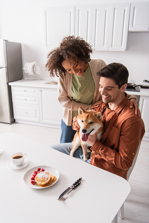 happy man holding shiba inu dog near breakfast and happy african american girlfriend