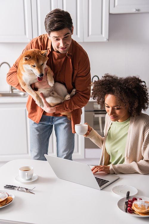 surprised man with shiba inu dog looking at laptop near african american girlfriend with coffee cup