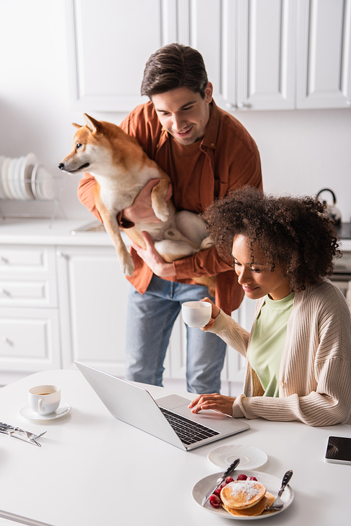 young man with shiba inu dog looking at laptop near smiling african american girlfriend