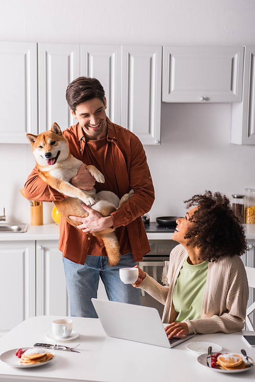 happy interracial couple looking at each other near shiba inu dog and laptop in kitchen