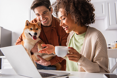 Smiling african american woman holding cup near devices and boyfriend with shiba inu at home