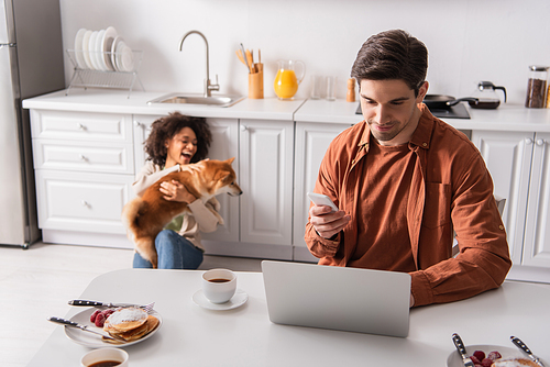 Man using smartphone and laptop near breakfast and african american girlfriend with shiba inu in kitchen