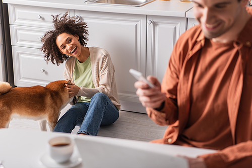 Smiling african american woman playing with shiba inu near blurred boyfriend with smartphone in kitchen
