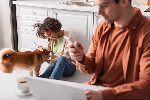 Young african american woman playing with shiba inu near boyfriend with devices in kitchen