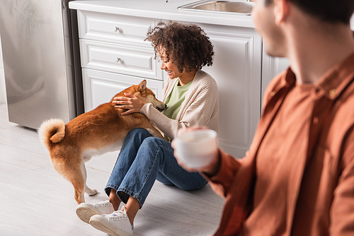 Young african american woman playing with shiba inu dog near blurred boyfriend with cup in kitchen
