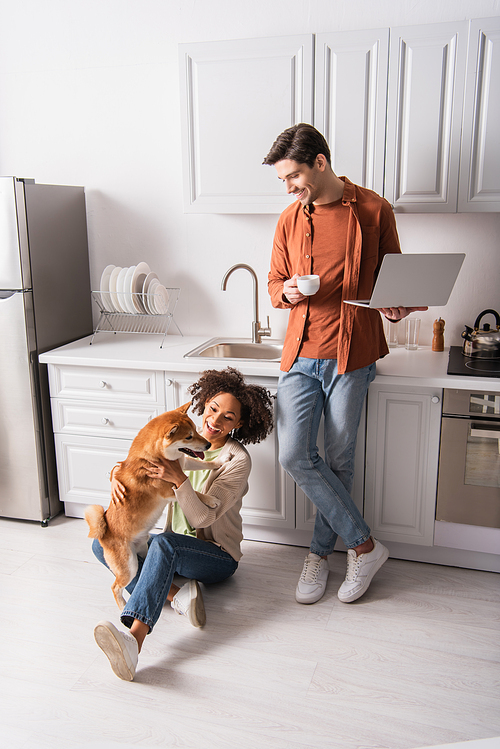Smiling man holding cup and laptop near african american girlfriend playing with shiba inu in kitchen