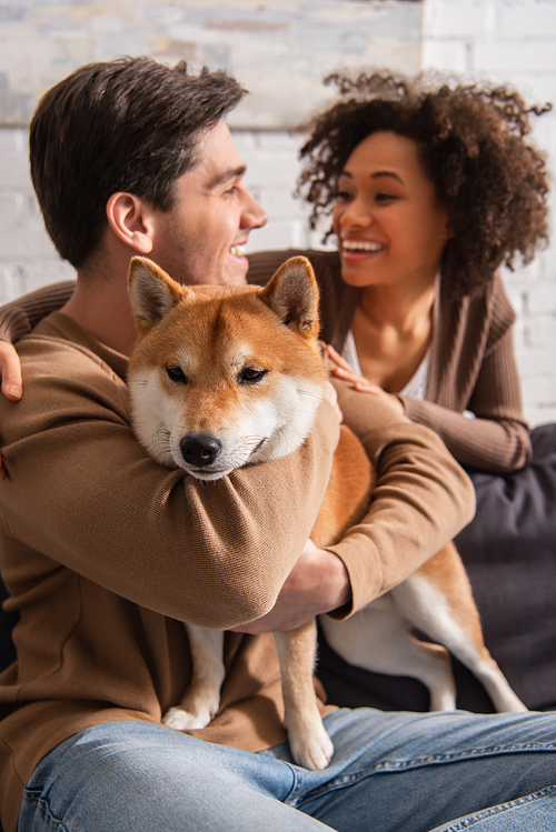 Smiling man hugging shiba inu dog near african american girlfriend at home