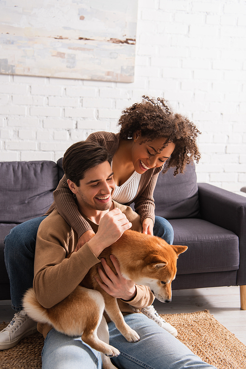 Smiling man petting shiba inu on floor near african american girlfriend on couch at home